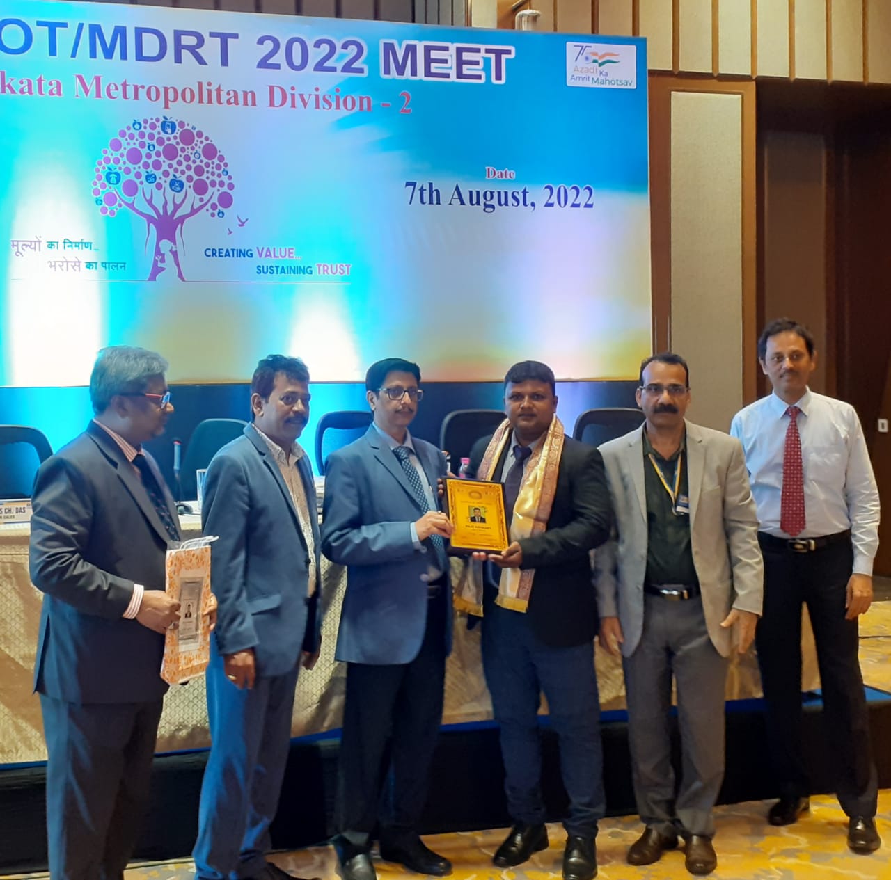 person writing in a notebook beside by an iPad, laptop, printed photos, spectacles, and a cup of coffee on a saucer