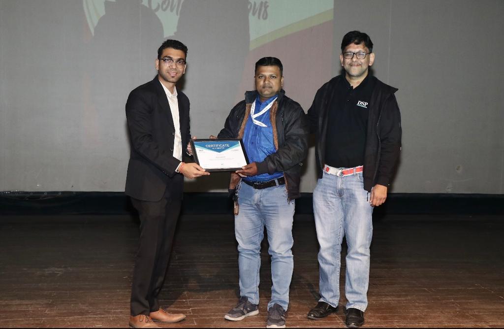 man wearing a black jacket, white shirt, blue jeans, and brown boots, playing a white electric guitar while sitting on an amp
